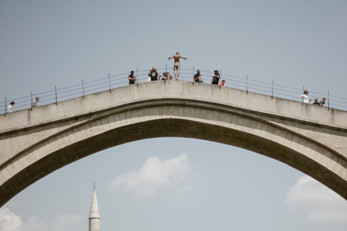 Bro i betong fotograferad snett underifrån med en svagt blå himmel som bakgrund. Några människor står på bron, en man i badbyxor ser ut att vara på väg att hoppa ner.  